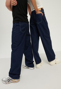 Zwei Personen stehen Rücken an Rücken, tragen dunkelblaue Jeans mit weitem Bein und lässige Sneakers, eine Hand in der Tasche, neutraler Hintergrund.