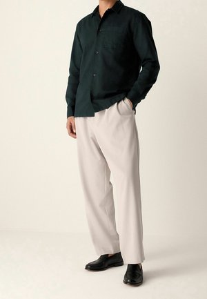 Mężczyzna w ciemnozielonej koszuli na guziki z długim rękawem, beżowych luźnych spodniach oraz czarnych skórzanych mokasynach, stoi z jedną ręką w kieszeni.