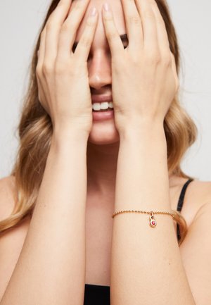 Mujer de cabello largo sonríe mientras se cubre el rostro con las manos, lleva una pulsera dorada con cuentas y un charm rojo en la muñeca derecha.