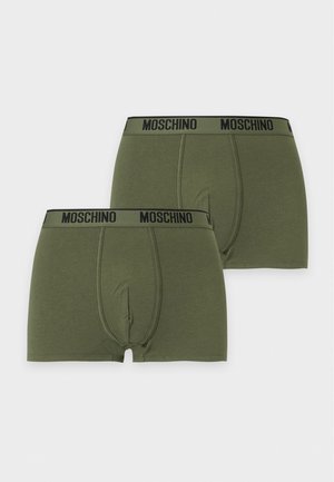 Un mannequin masculin se tient pieds nus devant un fond blanc uni, vêtu d’un boxer Moschino vert olive.