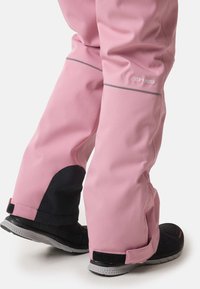 Lyserøde vandtætte bukser med grå detaljer og sorte strukturerede paneler ved anklerne. Har forstærkede ankler og justerbare fastlåsere.