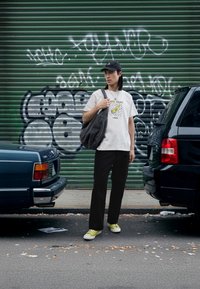 T-shirt blanc avec graphique et texte, pantalon noir, baskets vertes ; portant un sac fourre-tout sombre ; cadre urbain avec graffiti et voitures.