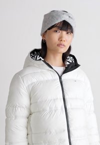 Giacca bianca imbottita con zip nera, indossata sopra un hoodie a righe bianco e nero. Accentuata da una fascia in maglia grigia. Texture liscia.