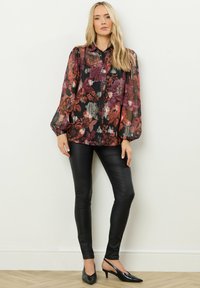 Bluse mit floralen Mustern aus transparentem schwarzem Stoff, in burgunder und gold akzentuiert, mit Kragen und puffärmeln, kombiniert mit schwarzen Lederleggings und Absatzschuhen.