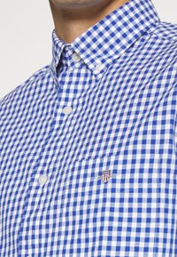 Blauw-witte ruitjesoverhemd met knoopsluiting op de kraag, voorzien van een borstzak en een geborduurd logo op de zak.