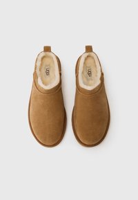 Bruine suède instapschoenen met een zachte bontvoering, voorzien van een ronde neus en een klein lusje aan de hiel. Gladde textuur.