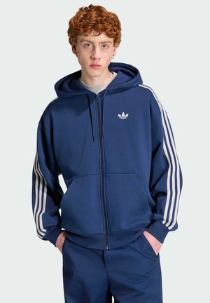 Młoda osoba z kręconymi rudymi włosami, ubrana w niebieską bluzę Adidas na zamek i pasujące spodnie, ręce w kieszeniach, neutralny wyraz twarzy.