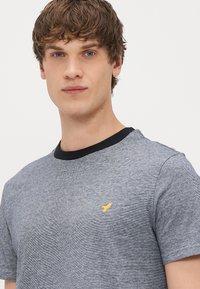 T-shirt gris chiné à manches courtes avec un col noir, arborant un petit logo d'oiseau jaune sur la poitrine. Tissu doux, coupe décontractée.