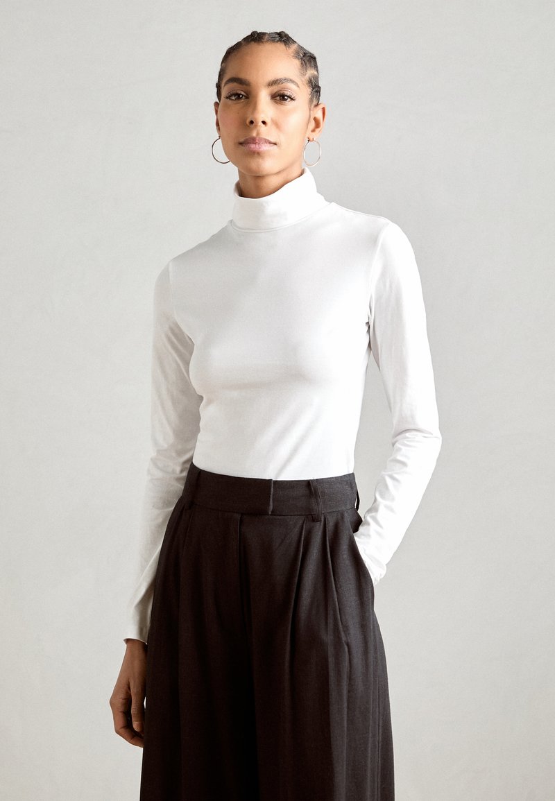 Top a maniche lunghe bianco con colletto a dolcevita abbinato a pantaloni neri plissettati a vita alta. Tessuto morbido, silhouette aderente e design minimal.