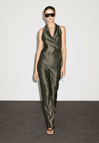 Olive green jumpsuit in satin fabric with a wrap design, featuring a v-neck and a cinched waist, paired with dark sunglasses and flat sandals.