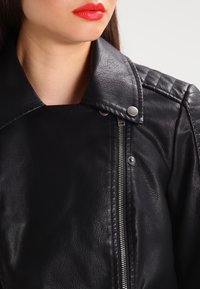 Kvinna i svart läderjacka med silverdragkedja och tryckknappar, synlig nedre del av ansiktet med röd läppstift, neutral bakgrund.