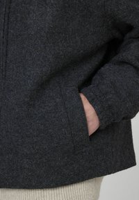 Veste en feutre gris charbon à coupe décontractée, avec une poche avant et un pull en tricot beige clair en dessous. Texture douce.