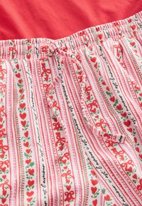 Spodnie z elastycznym paskiem w talii, wykonane z białej tkaniny zdobionej czerwonymi sercami, kokardkami, zielonymi winoroślami oraz czarnym francuskim pismem, związane pasującym sznurkiem.