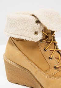 Botte compensée couleur fauve avec manchette en peau lainée rabattue et lacets jaune-marron passant à travers des œillets métalliques sur fond blanc.