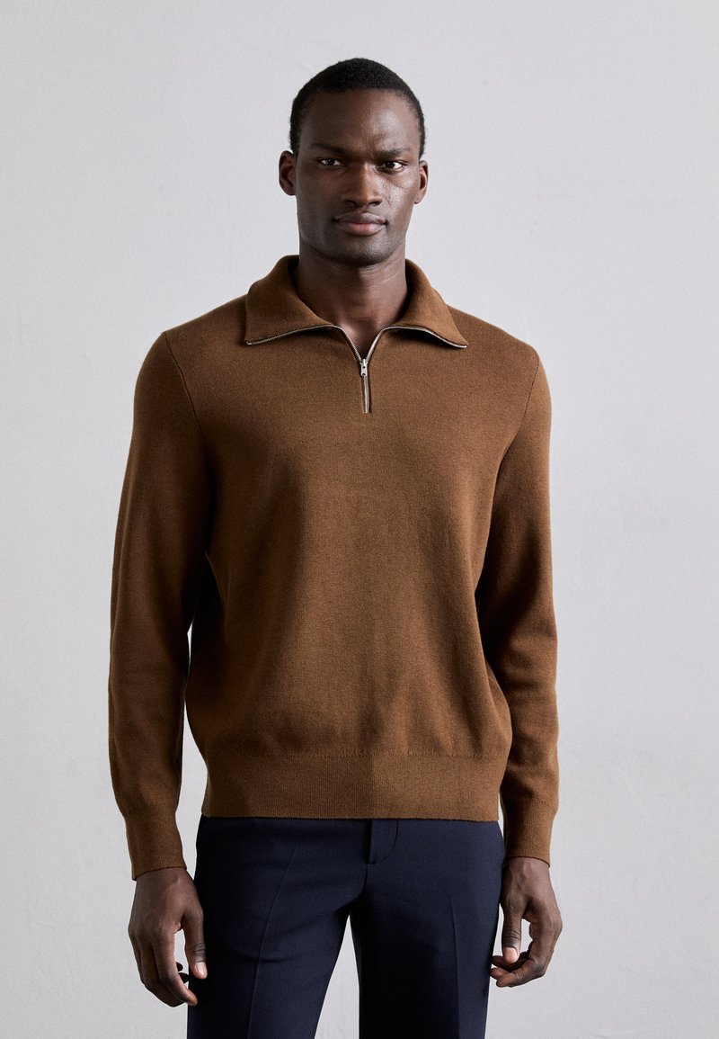 Pull à col zippé marron en tricot doux, avec un col plat et des poignets côtelés, associé à un pantalon noir cintré.