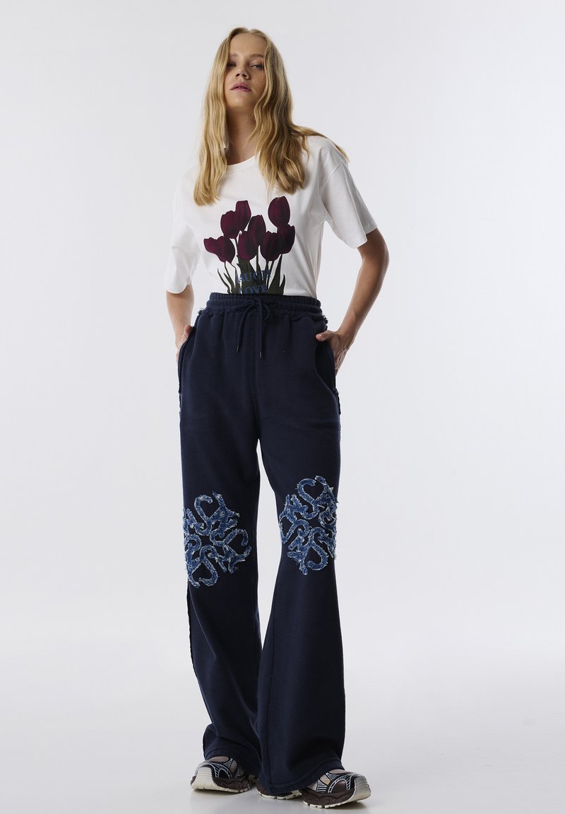 Marineblauwe sweatpants met geborduurde bloemmotieven op beide benen; gecombineerd met een wit T-shirt met een bloemenpatroon. Sneakers maken de look af.