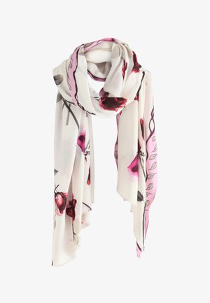 Kvinde iført beige trenchcoat, grå sweater, sorte bukser, sorte sko og hvidt tørklæde med pink blomsterprint, står med hænderne i lommerne.