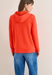 Femme portant un sweat à capuche orange et un jean bleu, debout de profil avec le dos partiellement tourné vers la caméra sur un fond beige.