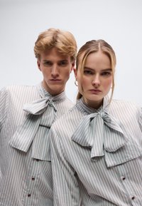 Οι ριγε μπλούζες με κουμπιά έχουν ψηλό γιακά με φιογκάκι στη λαιμόκοψη. Η παλέτα χρωμάτων περιλαμβάνει ανοιχτό μπλε, λευκό και διακριτικές καφέ ρίγες.