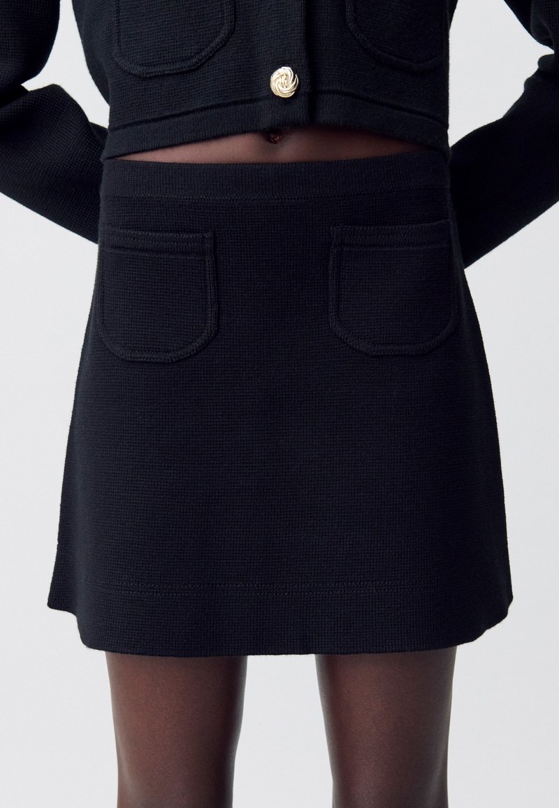 Jupe en tricot noir avec une finition texturée, dotée de deux poches avant et d'un léger évasement à l'ourlet. Accentuée par un détail en métal doré.