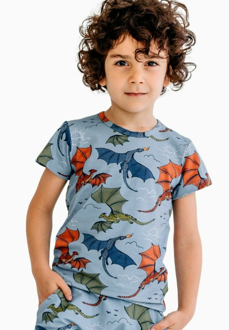 Young child with curly hair wearing matching blue pajamas covered in colorful flying dragons, standing with hands in pockets.