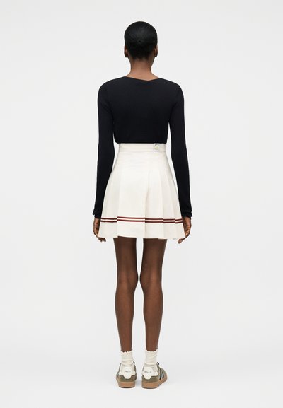 Γυναίκα που στέκεται με την πλάτη στραμμένη, φοράει μαύρο μακρυμάνικο τοπ, λευκή πλισέ φούστα με δύο κόκκινες ρίγες κοντά στο τελείωμα, λευκές κάλτσες και αθλητικά παπούτσια.