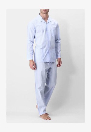 Uomo in piedi a piedi nudi che indossa una camicia azzurra chiara a maniche lunghe con bottoni e pantaloni ampi abbinati su sfondo semplice.