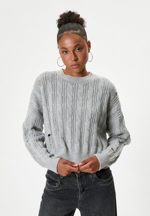 Grijze kabelgebreide sweater met een ronde hals, geribde manchetten en een zichtbare naaddetail. Gecombineerd met hoge zwarte jeans.