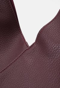 Sac en cuir bordeaux avec une surface texturée, une finition lisse et des coutures rouges contrastées le long des bords structurés.
