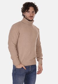 Maglione beige a collo alto con un motivo a maglia strutturato, polsini e orlo a costine, indossato con jeans di denim blu. Design semplice e classico.