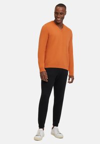 Maglione arancione a V in maglia, materiale testurizzato, abbinato a pantaloni neri tapered e sneakers bianche. Nessun motivo o decorazione visibile.
