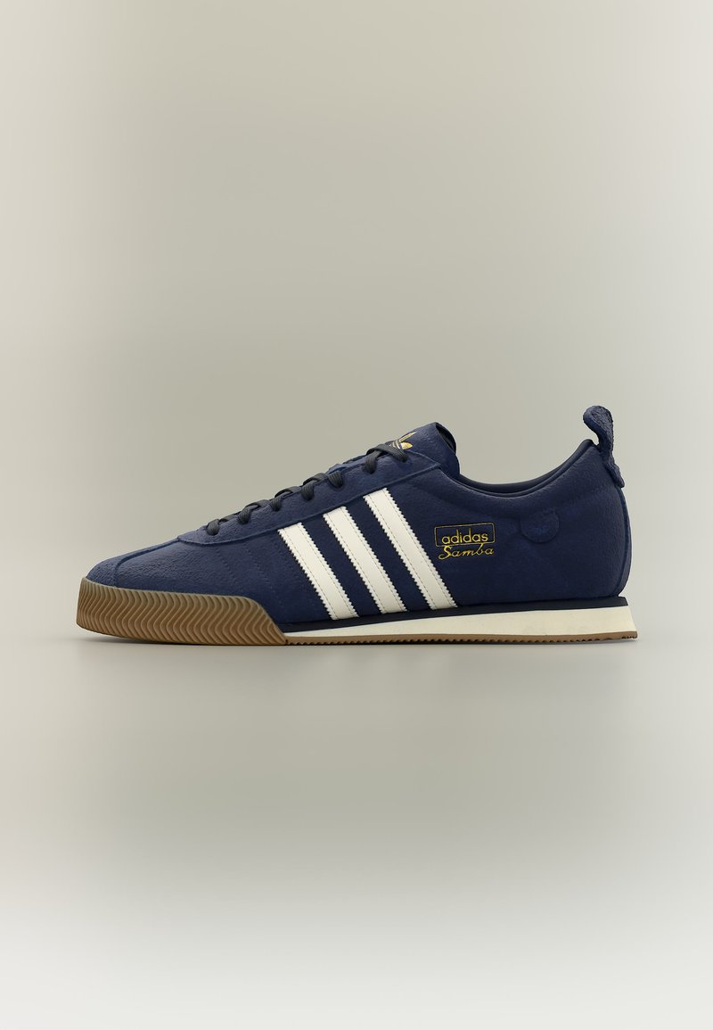 Baskets Adidas Samba bleu marine avec des rayures blanches, semelle en gomme et étiquette dorée « adidas Samba » sur le côté.
