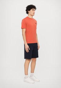 Oransje bomull T-skjorte med korte ermer og en liten svart logo på brystet, sammen med marineblå shorts og hvite sneakers.