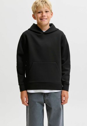 Garçon blond souriant portant un sweat à capuche noir sur une chemise blanche et un pantalon gris, debout devant un fond clair uni.