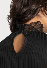 Svart ribbad topp med en spetskrage med vågiga kanter och tre svarta knappar baktill, som framhäver en nyckelhålssiluett.