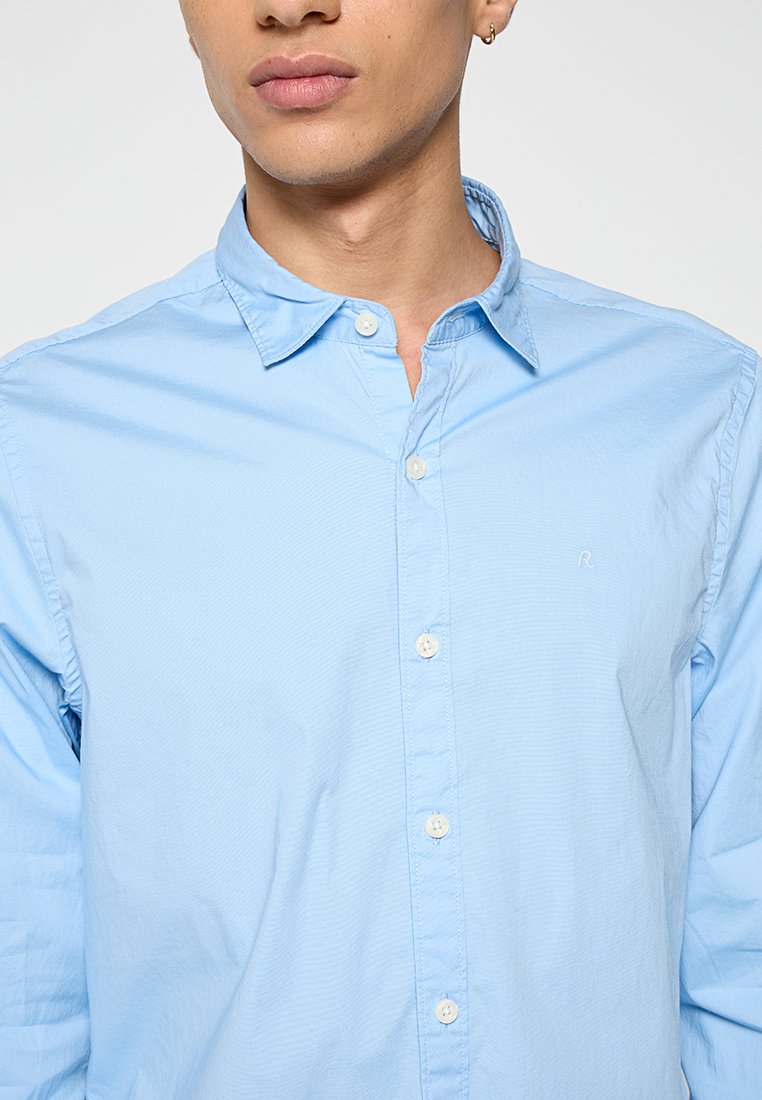 Camicia button-up azzurro chiaro in cotone, caratterizzata da un colletto classico, un tessuto testurizzato e un logo discreto sul lato sinistro.