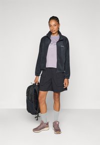 Giacca di felpa grigia scura, maglietta lavanda a maniche corte, pantaloni corti neri, calzini grigi e scarpe marroni. Zaino tenuto nella mano sinistra, look casual.