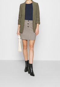 Blazer vert olive porté sur un haut bleu marine, assorti à une jupe à motif pied-de-poule avec des boutons noirs. Bottines noires et sac crème.