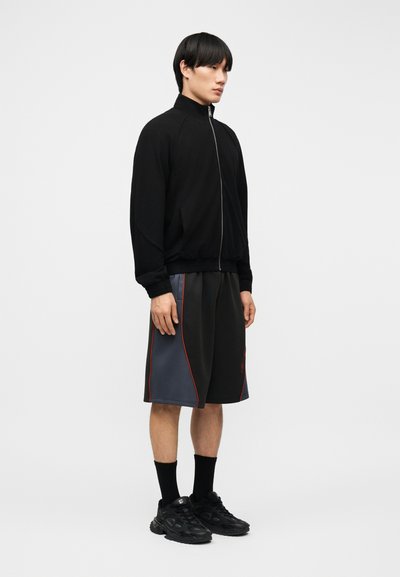 Νεαρός άνδρας που στέκεται φορώντας μαύρο μπουφάν με φερμουάρ, μαύρο και γκρι σορτς με κόκκινη ρίγα, μαύρες κάλτσες και μαύρα αθλητικά παπούτσια, μπροστά σε λευκό απλό φόντο.