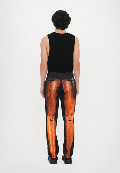 Schwarzes ärmelloses Oberteil mit figurbetontem Design, kombiniert mit schwarzen Hosen, die auf der Rückseite mit einem Holz-Muster bedruckt sind und mit Taschen versehen sind.