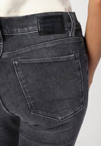 Jeans de mezclilla gris oscuro con un acabado texturizado, con un parche de cuero negro con la marca en la cinturilla y sutiles detalles de costura.