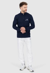 Pullover navy a maniche lunghe con zip a quarto e logo, abbinato a pantaloni bianchi e scarpe da ginnastica, con una vestibilità casual e una texture liscia.