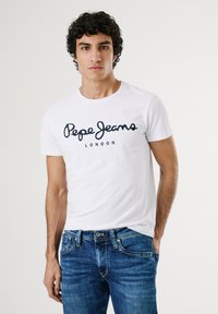 Bijela pamučna majica s crnim logom "Pepe Jeans" i natpisom "LONDON" ispod. Model nosi plave traperice. Jednostavni okrugli izrez i kratki rukavi.