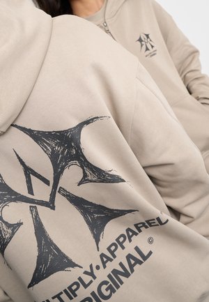 Dwie osoby w beżowych bluzach z kapturem z czarnym graficznym wzorem krzyża i tekstem "Multiply Apparel Original" widocznym na materiale.