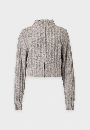 Une femme porte un cardigan en maille gris, un haut blanc, un pantalon noir évasé et des escarpins pointus sur un fond de studio uni.