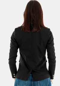 Blazer nero con un design aderente, tessuto strutturato e dettagli in bottone dorato sui polsini. La parte posteriore presenta una fessura centrale e linee morbide.