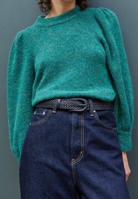 Kraftig grön stickad tröja med puffärmar, tillsammans med högmidjade mörka denimjeans och ett flätat svart bälte med en rund spänne.