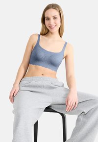 Modrá braletka s textúrovaným vzorom, štvorcovým výstrihom a širokými ramienkami; skombinovaná so sivými teplákmi, sediaca na čiernej stoličke.