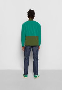 Jakke i grønne og olivengrønne farveblokke lavet af blødt stof, med høj krave og afslappet pasform, kombineret med mørkeblå jeans og grønne sneakers.