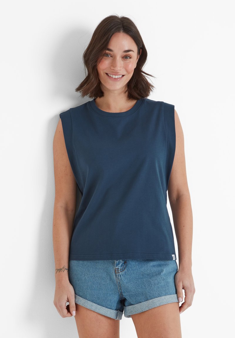 Woman with shoulder-length dark hair wearing sleeveless navy blue top and rolled-up denim shorts, smiling against white background